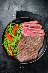Grilled medium rare flank beef steak with salad in a plate. Black background. Top view