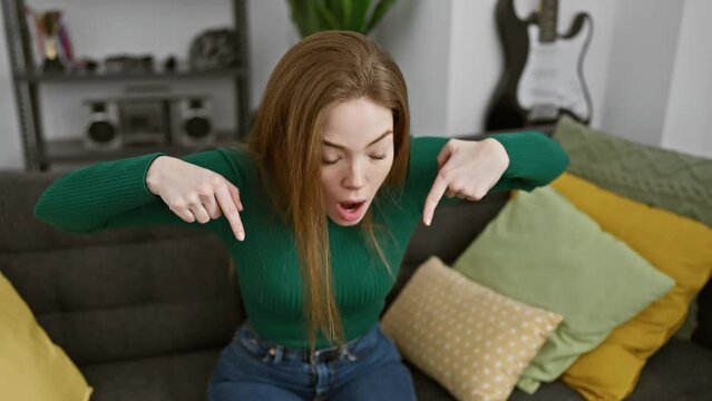 Surprised young blonde woman at home, pointing down in awe - showing ad with a wow-factor wearing a cozy sweater. beautiful surprise expression on her face!