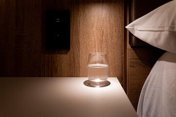 Small side table next to a hotel bed illuminated by a directional lamp with a glass of fresh water