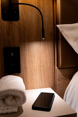 Small table next to luxury hotel bed illuminated by directional lamp with mobile phone and rolled towel