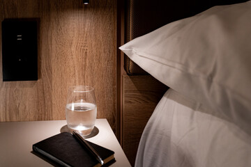 Small table next to bed in hotel room illuminated by small lamp with agenda pen glass of water and...