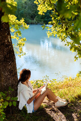 Young woman writing and drawing in notebook sitting under the tree near river on nature in sunlight. Closeness to nature  inspires creativity. Urban escapism.