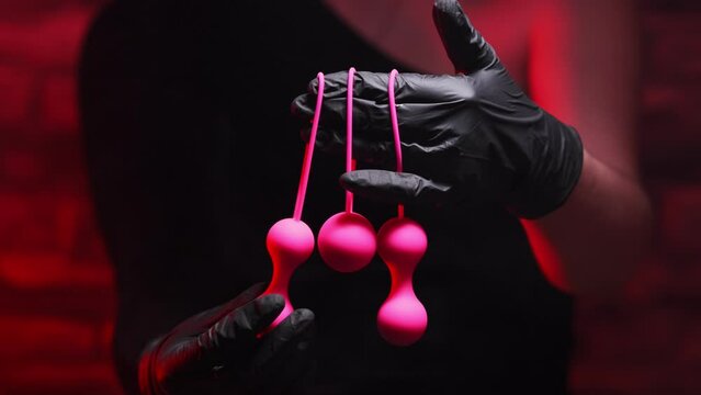 Woman's hand in black latex gloves and a black dress holds pink Kegel balls and Ben Wa balls on a red background. Vaginal balls in a woman's hand. Sex toys Geisha Balls, Products for intimate stores
