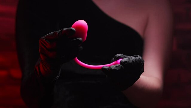 Pink vaginal vibrator for the G-spot in the hand of a girl in black latex gloves and a black dress on a red background. Sex Shop Products, Adult Gifts for Couples, Adult Store
