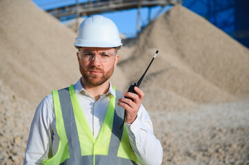 A worker in a hard hat stands with a walkie-talkie at a stone crushing plant.