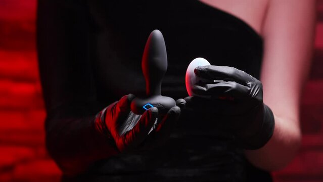 Silicone anal plug with a remote control rotates in a female hand in black latex gloves and a black dress on a black background. Massager with multiple speeds. Sex shop products, adult gifts
