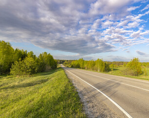 Asphalt road through a field, Evening landscape. Blue sky with clouds above the horizon. early spring in the countryside, soft sunlight on the grass.