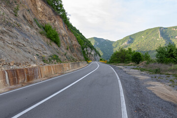  winding road that cuts through a mountainous landscape. The road is devoid of traffic, with guardrails and landslide netting visible, indicating recent construction or maintenance.