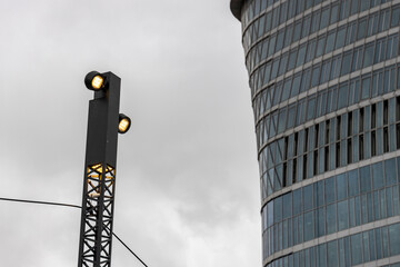 Obraz premium A modern streetlight with illuminated light bulbs stands against the backdrop of a towering skyscraper with a reflective glass facade, under an overcast sky in the city center.