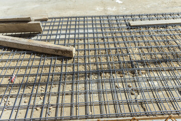 A close-up of a construction site with a grid of rebar laid out on the ground. A concrete block and formwork are also visible, indicating the preparation of a concrete slab or foundation.