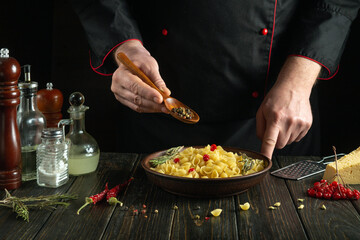 An experienced chef uses a spoon to add dry pepper to a bowl of pasta on the kitchen table. The concept of preparing a delicious breakfast in a restaurant kitchen.