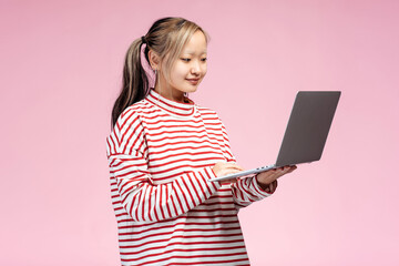 Happy cute Asian student using laptop looking at screen, isolated on pink background