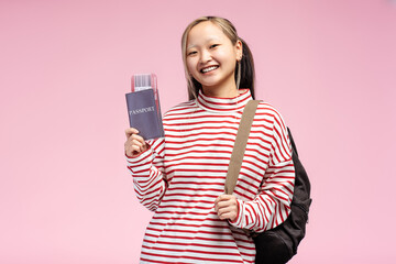 Happy Asian female student holding passport with plane ticket and backpack on pink background