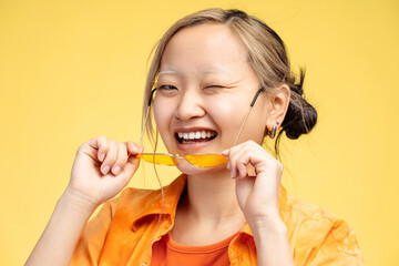 Happy pretty Asian girl holding stylish glasses looking at camera, isolated on yellow