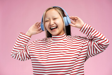 Expressive Asian girl singing song wearing headphones posing in studio, isolated on pink