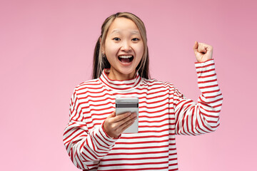 Excited Asian girl posing and looking at camera using mobile phone, isolated on pink