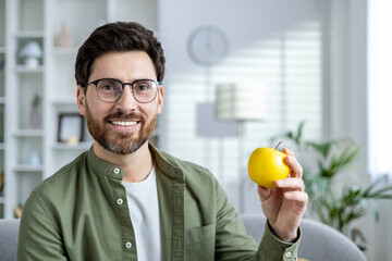 A cheerful man with glasses holding a yellow apple, sitting in a bright, modern living room. The...