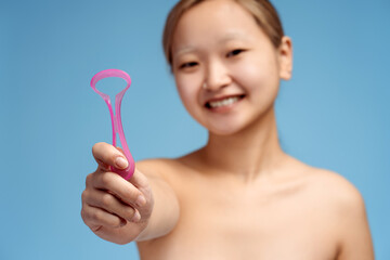 Happy Asian girl holding device for hygienic cleaning for tongue, isolated on blue