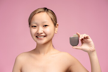 Smiling Asian girl with bare shoulders showing pacemaker in hand, isolated on pink