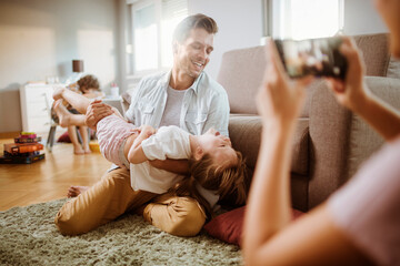 Joyful father playing with daughter while mother takes photo with smartphone at home