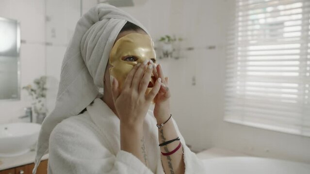 Medium shot of Caucasian woman applying golden sheet mask on face during skin care procedure at home