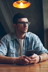 Concentrated young hipster guy pondering looking away holding telephone device in hand and waiting important call via application, thoughtful man in spectacles for vision correction using technology