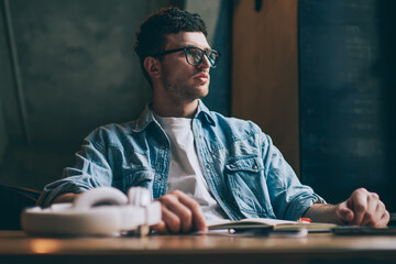 Pondering caucasian hipster guy sitting at desktop and thinking about studying indoors, pensive young man in spectacles for provide eyes protection thoughtful looking away while waiting friend