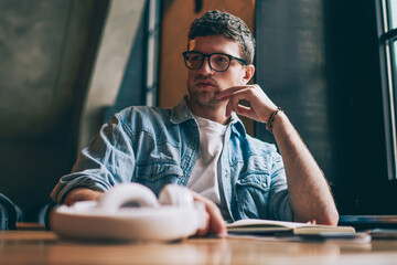 Thoughtful young hipster guy in eyewear pondering on organisation plan sitting at desktop...