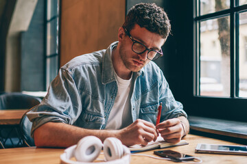 Serious male writer sitting at desktop with textbook for education and holding pen in hand while thinking on information for article, pensive hipster guy in eyeglasses enjoying autodidact indoors