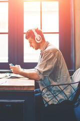 Young caucasian hipster guy playing games via mobile application while listening motivation audio book via headphones indoors, man enjoying free time on leisure with music and smartphone device