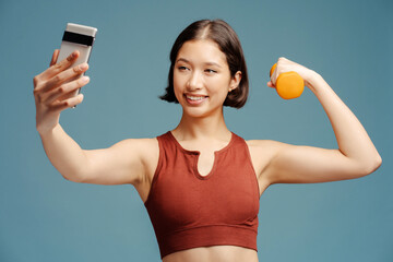 Smiling woman holding mobile phone and dumbbell, making selfie isolated