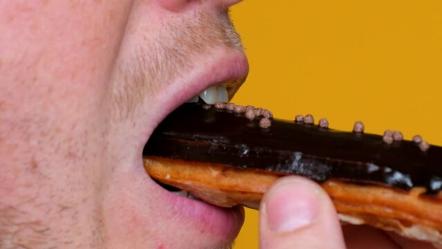 Close-up side view of a man's unshaven face, mouth bites off a piece of eclair cake and chewing. on a yellow isolated male background. bright tasty delicious pastry, the concept junk food