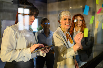 Female mature manager writing on sticky notes and giving tasks to team