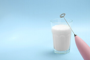 Mini mixer (milk frother) and whipped milk in glass on light blue background. Space for text