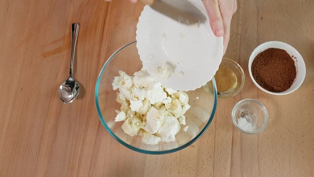 The chef prepares a healthy banana dessert. Add ricotta, cocoa, vanilla and honey to a glass bowl with sliced ​​banana. Close-up, top view