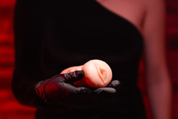 Sex toys for men. An artificial vagina made of silicone for masturbation in the hands of a girl in black gloves and a black dress on a red background. Sex shop Adult store