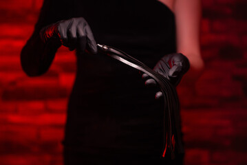 Close-up of female hands in black gloves holding a BDSM whip on a red background. BDSM is an outfit...