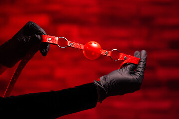 Close-up of female hands in black latex gloves holding a red BDSM gag on a red background. BDSM -...