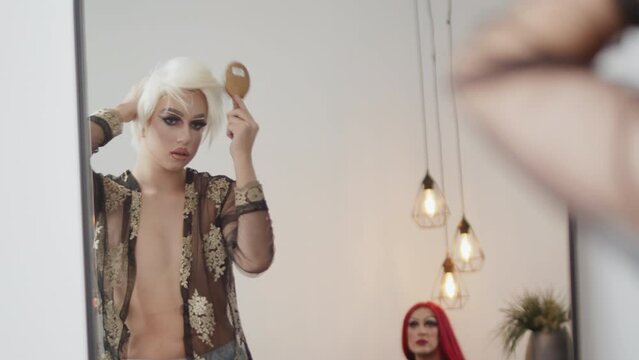 Young performer in a drag queen makeup looking in a mirror and combing his wig with a comb in the dressing room during preparation for the show