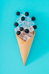 Ice cream cone with blueberries and ice cubes on aqua blue background. Minimal summer creative layout. Fruit and candy concept. Summer sweet food aesthetic. Flat lay.
