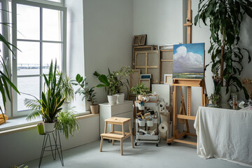 Cozy creative artist workplace with easel, oil artwork painted on canvas. Painter home art studio decorated with multiple green fresh plants, potted flowers. Inspiring bright work environment