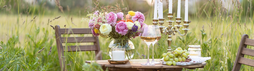 Elegant wedding or private party for a loving couple table setting outdoors in the garden. Beautiful decor with fresh ranunculi flowers, candles. Gourmet cheese board, fruits, white wine. Banner