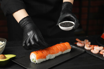 Chef in gloves adding sesame seeds onto tasty sushi rolls at black wooden table, closeup - Powered by Adobe
