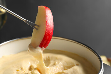 Dipping piece of apple into fondue pot with melted cheese on grey background, closeup