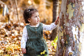 Fashionable cute little standing among golden autumn leaves and touching huge tree