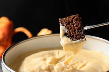 Dipping piece of bread into fondue pot with melted cheese on black background, closeup