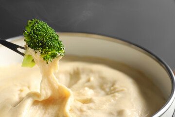 Dipping piece of broccoli into fondue pot with melted cheese on grey background, closeup