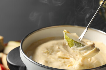 Dipping piece of pear into fondue pot with melted cheese on grey background, closeup