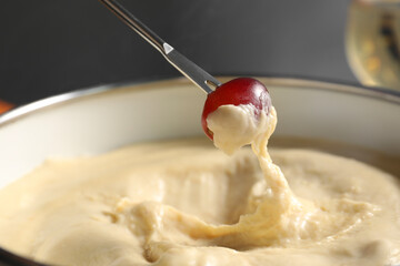 Dipping piece of grape into fondue pot with melted cheese on grey background, closeup