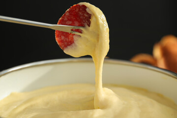 Dipping piece of sausage into fondue pot with melted cheese on black background, closeup